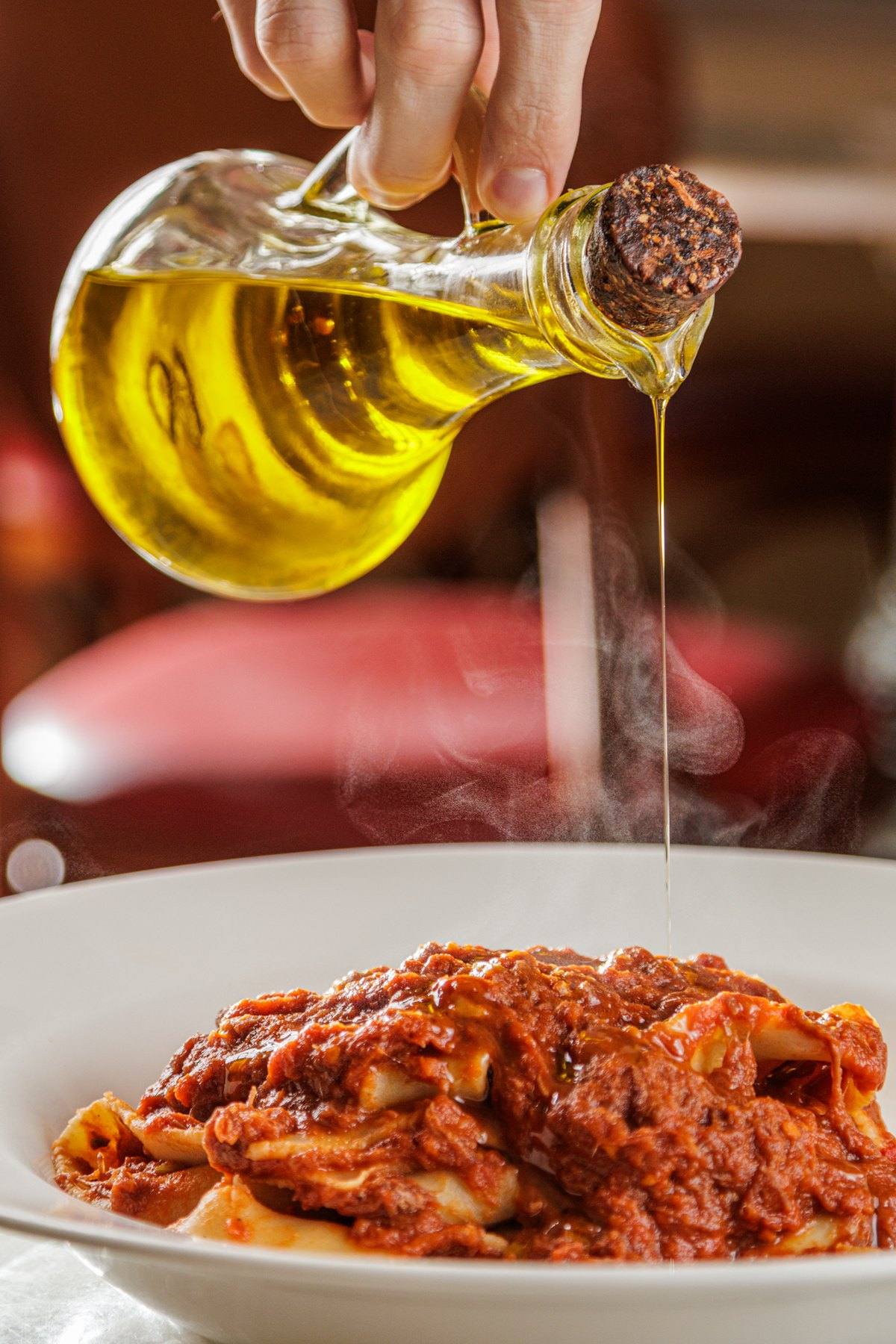 Azeite sendo servido sobre um prato de pappardelle ao ragù de ossobuco, com as cadeiras vermelhas do salão ao fundo